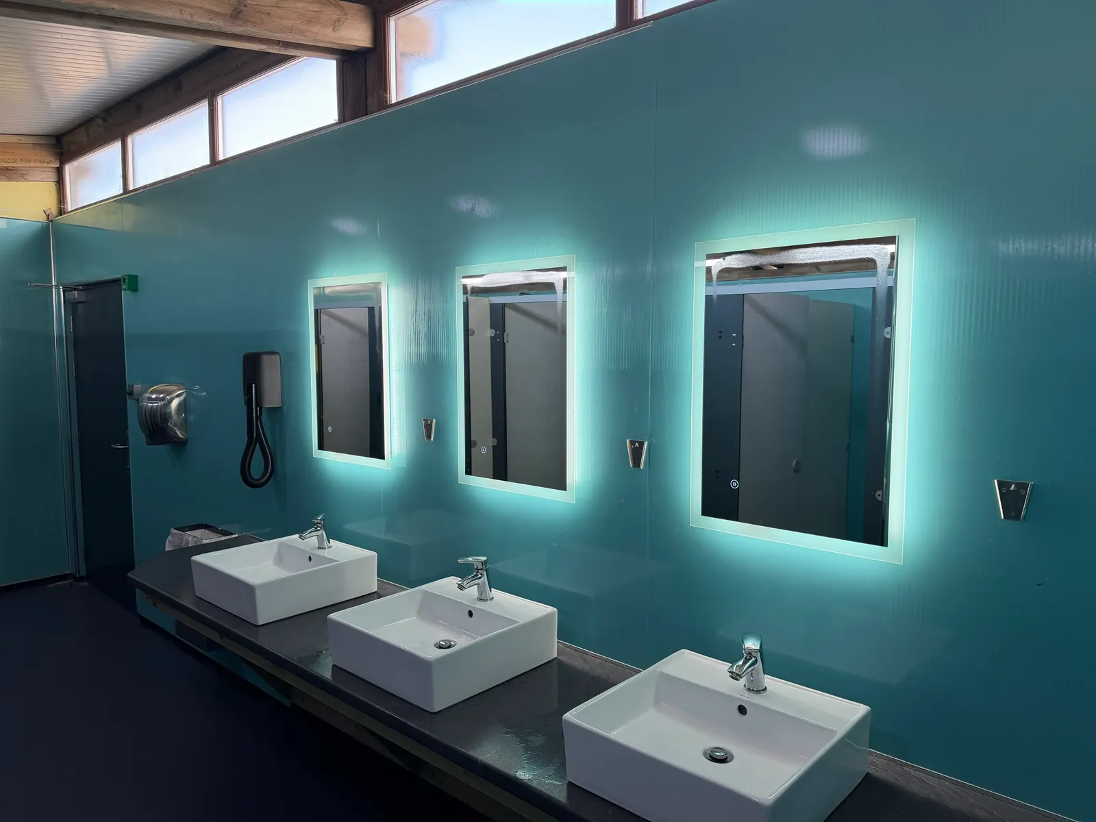 Three white basins under lit mirrors, with a hairdryer and a hand dryer on the wall.