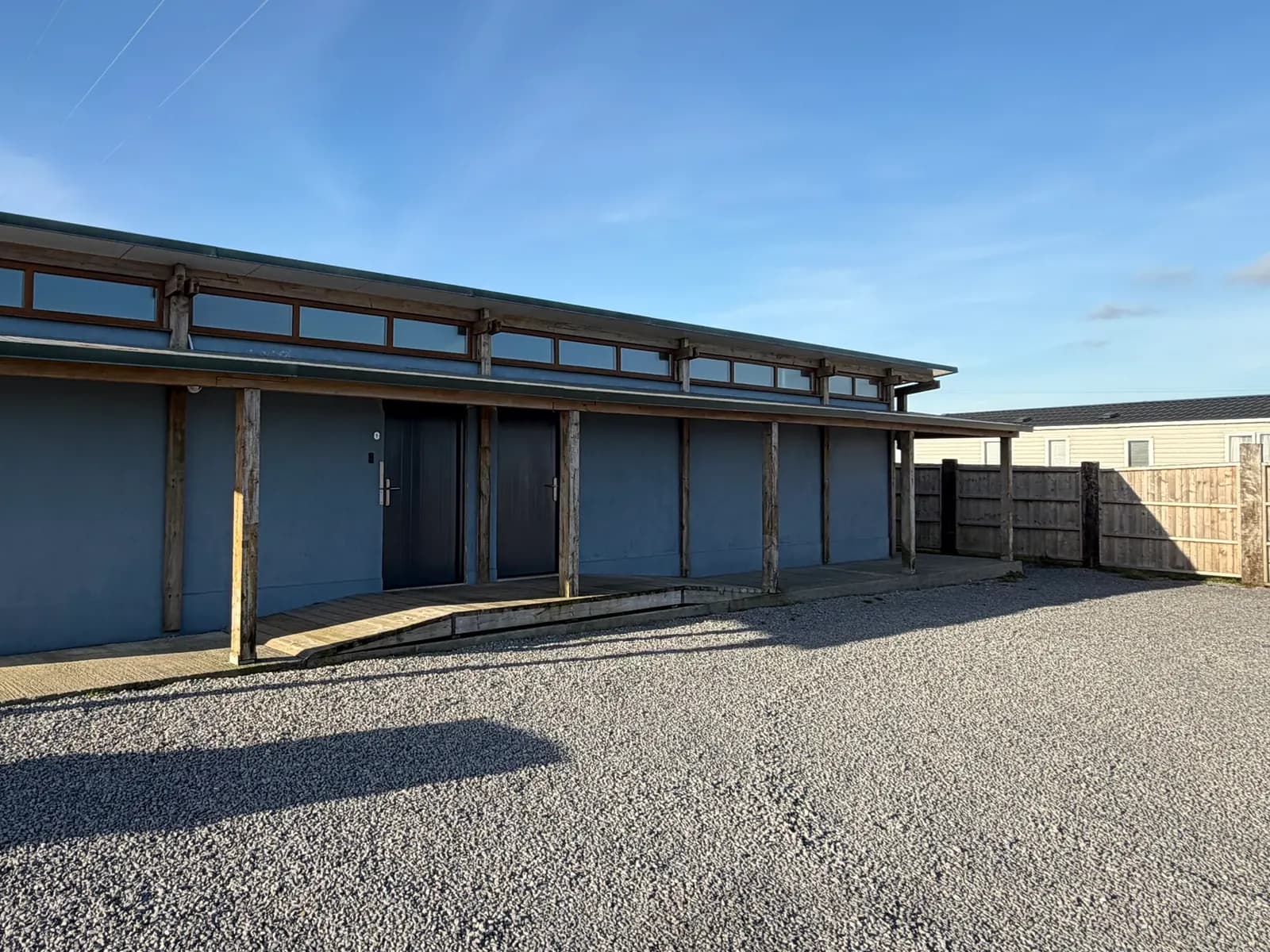 The toilet and shower block from outside: a long blue building with a covered walkway and a wooden ramp up to the doors, beside a gravel yard.
