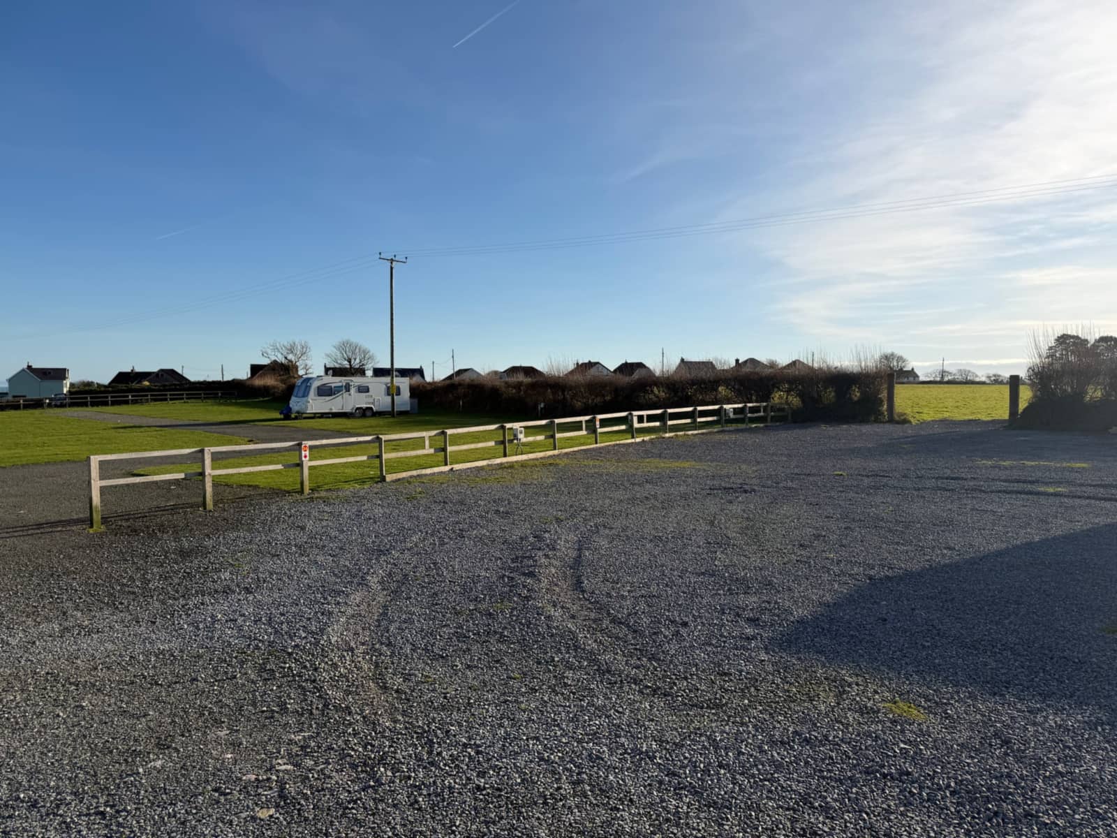 The hardstanding area at Broadfield: a wide, open gravel area with a wooden rail fence along one side, electric hook-up points and a pitch number on the fence, and the grass camping field behind it.
