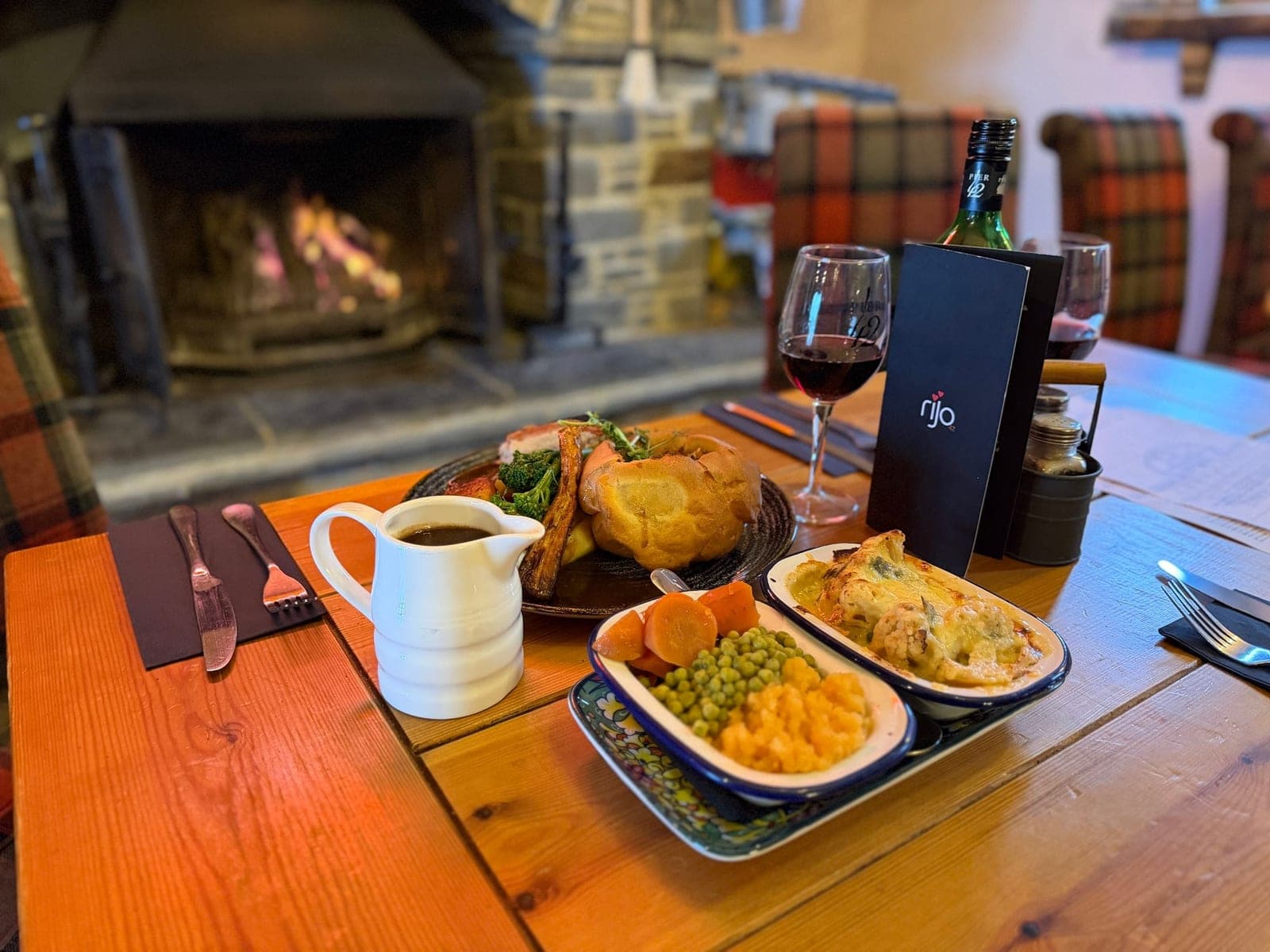 A Sunday roast at Jan'z by the open fire: a plate with a Yorkshire pudding, a jug of gravy, dishes of vegetables and cauliflower cheese, and a glass of red wine.