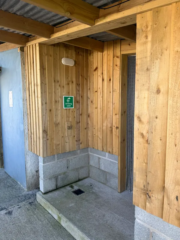 The drinking water tap on a wooden wall under a roof, with a green sign above it saying Drinking water.