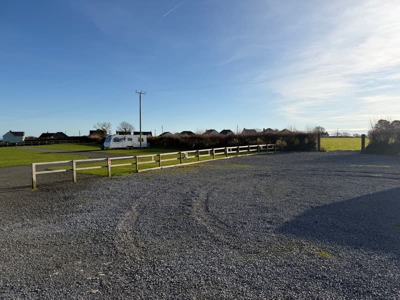 The hardstanding area at Broadfield: a wide, open gravel area with a wooden rail fence along one side, electric hook-up points and a pitch number on the fence, and the grass camping field behind it.